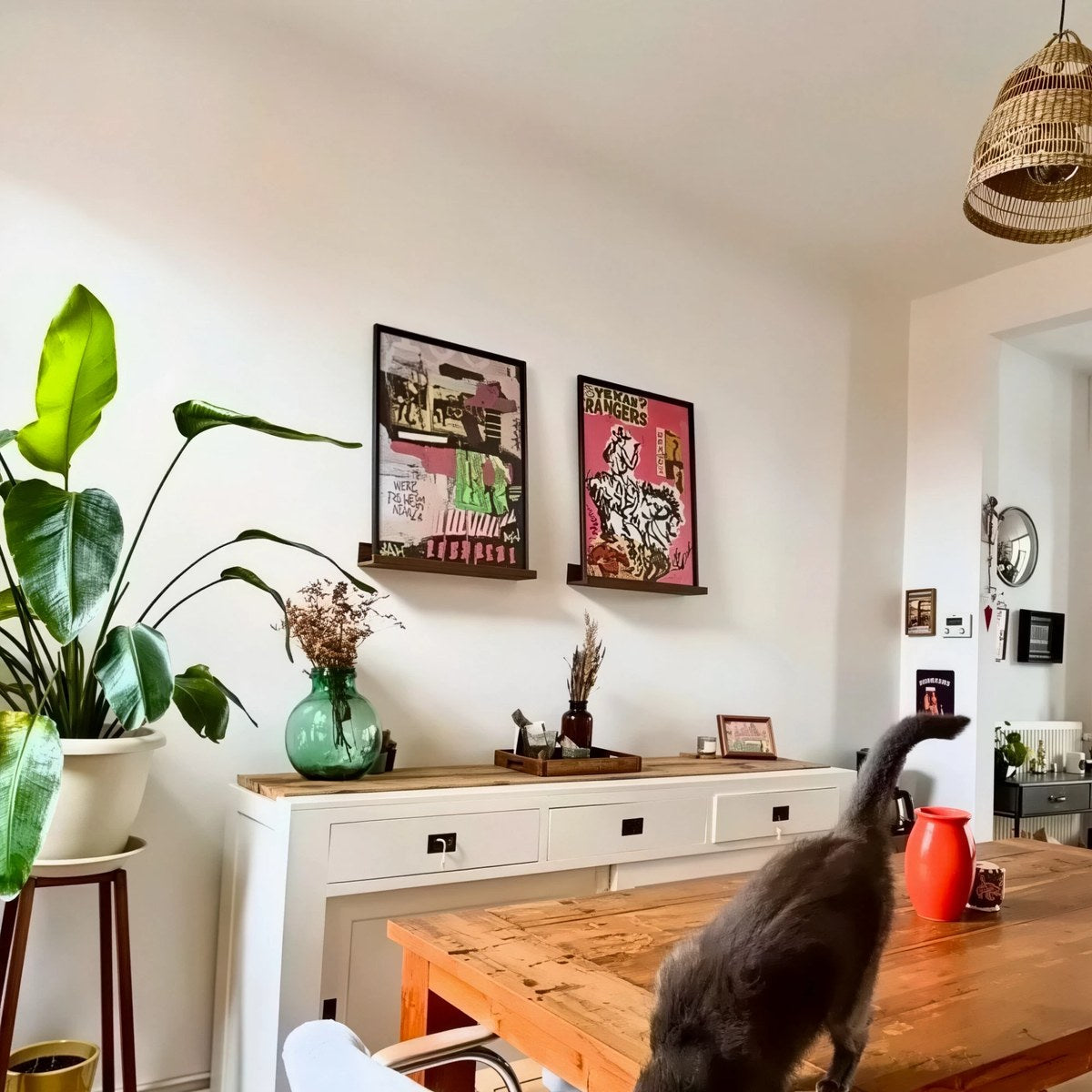 Dos pósters personalizados con marco negro sobre repisas de madera, encima de un aparador blanco, en un comedor con una planta y un gato